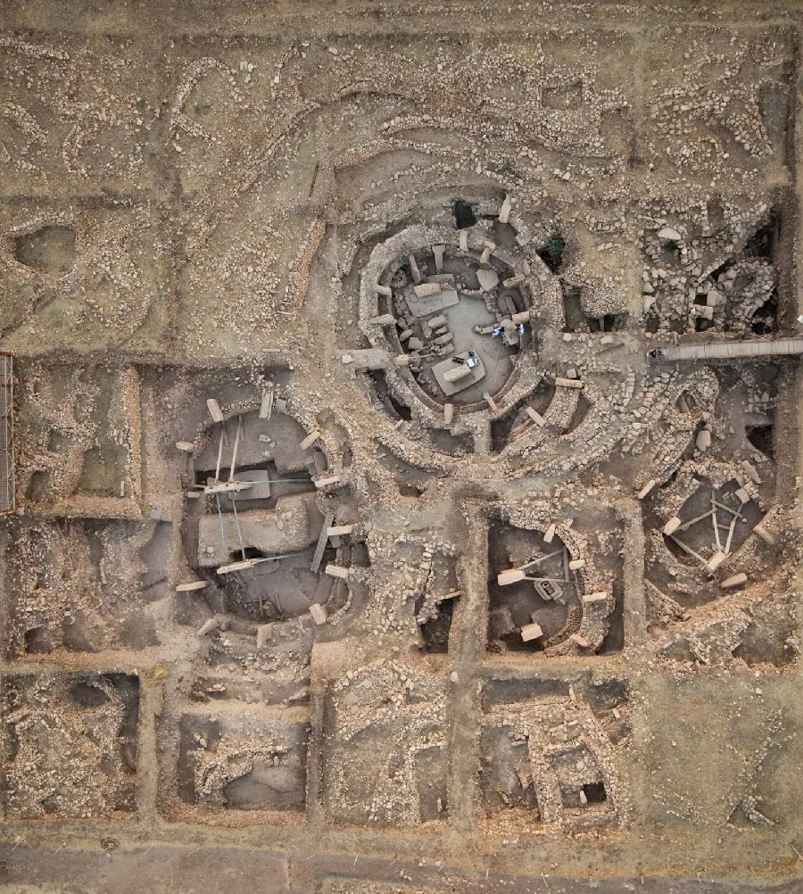 An aerial photo is shown of an old structure made up of stones and dirt. There are four circular areas throughout the picture and one square area with stones in various shapes arranged inside the areas, some on the ground and some stacked on each other. Rectangular, large stones are arranged around the perimeters of those areas, lighter in color than the rest. Around the perimeter of the entire area, there are square areas, mostly filled with dirt, showing the walls of the areas sticking up from the ground. One square area toward the top right of the picture is filled with more stones and is dug deeper than the rest.