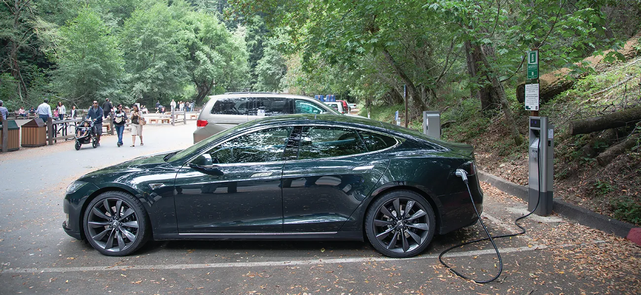 A photograph is shown of a parked car plugged into a charging station in a paved parking area. The parking area is situated in a wooded area. People are walking in the background in the park-like atmosphere.