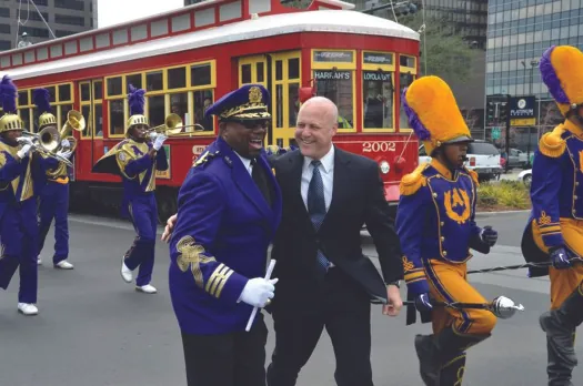 An image of Mitch Landrieu standing in the middle of a group of people who are playing various instruments. A streetcar is in the background.