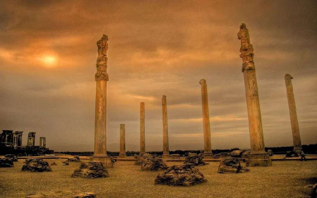 A picture of seven broken, brownish columns at sunset is shown. Some of the columns have decorative pieces at the top. Pedestals litter the ground. In the left background are broken double columns atop a large dark platform. The ground is gravelly and trees can be seen in the far background.