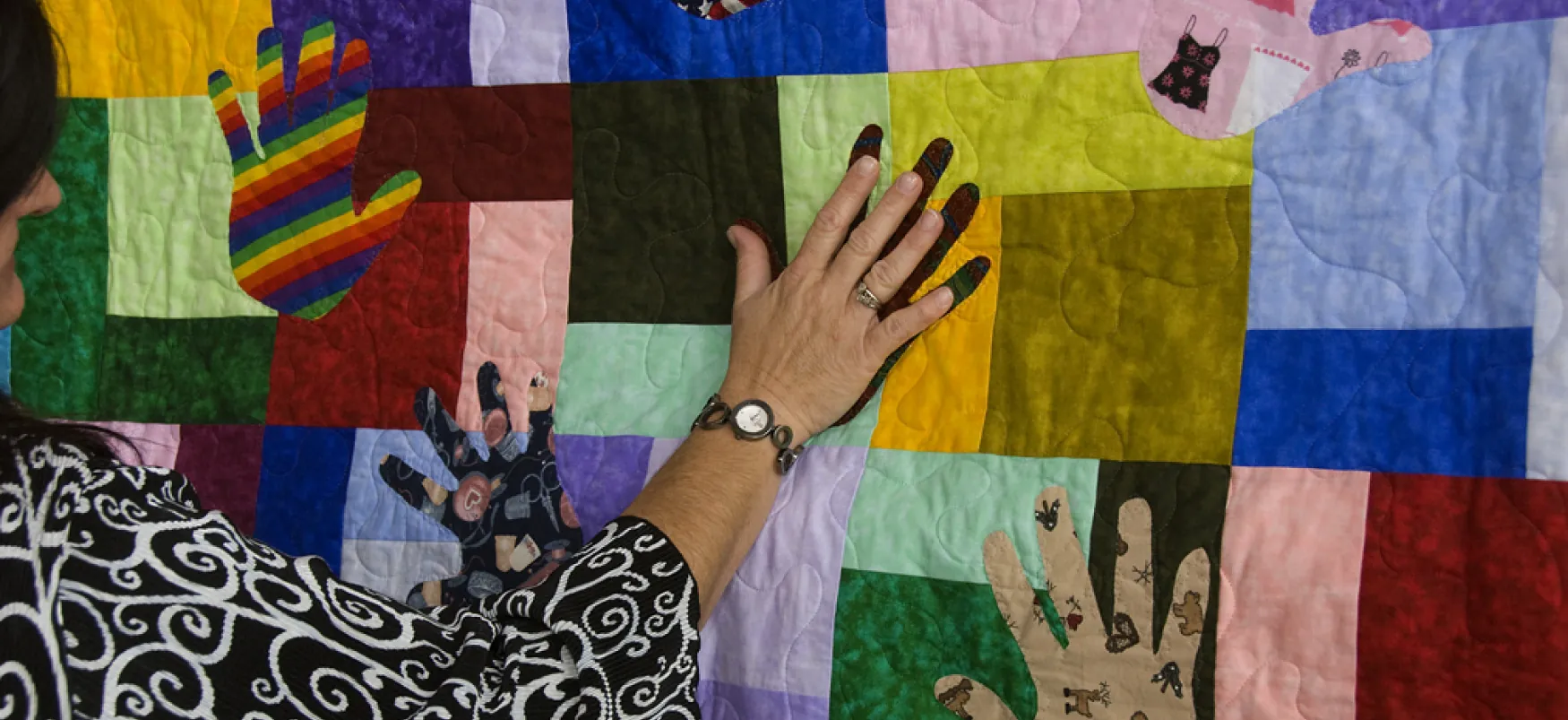 A hand touches a patchwork quilt. In addition to the traditional squares, the quilt has hand-shaped pieces.