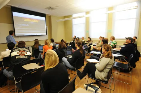Teacher and high school students in a classroom looking at the projection screen in the front of the classroom.