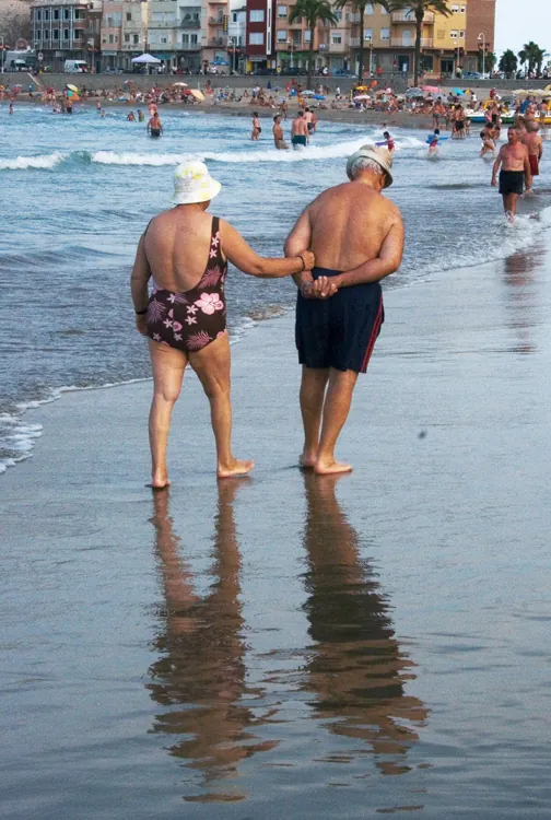 An elderly man wearing blue swim trunks and an elderly woman wearing a flowered bathing suit and hat are shown walking near the water on a beach.