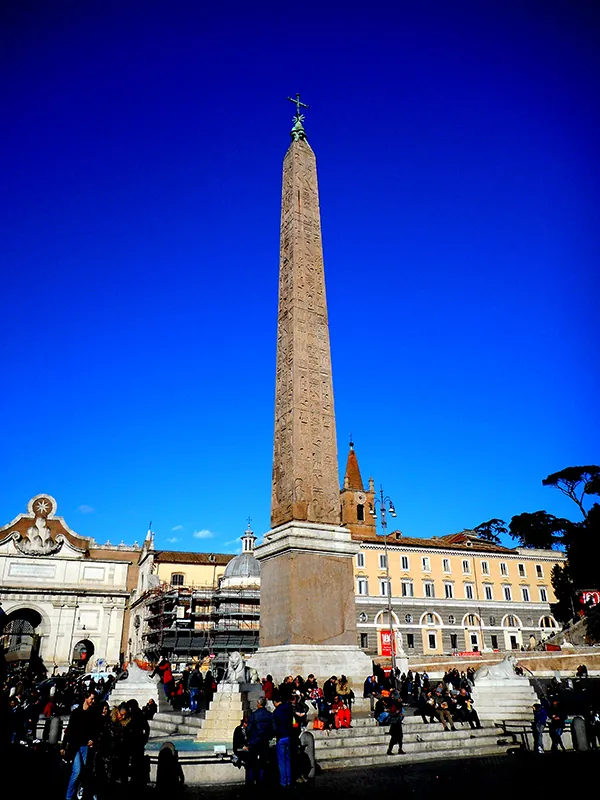 Very tall Egyptian obelisk in the middle of a public square. Behind it are several four-story stone buildings. People sit at the obelisk’s base.