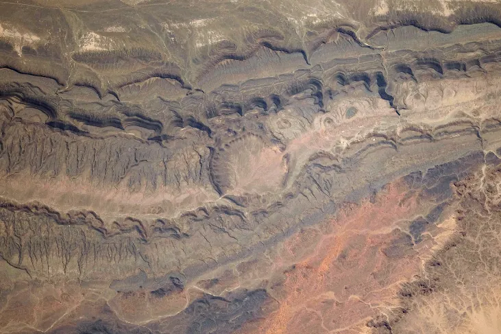 Photograph of an Impact Crater from Space. The large, circular Ouarkziz crater clearly stands out in the center of this image amidst the parallel lines of the mountains and ridges where it lies.