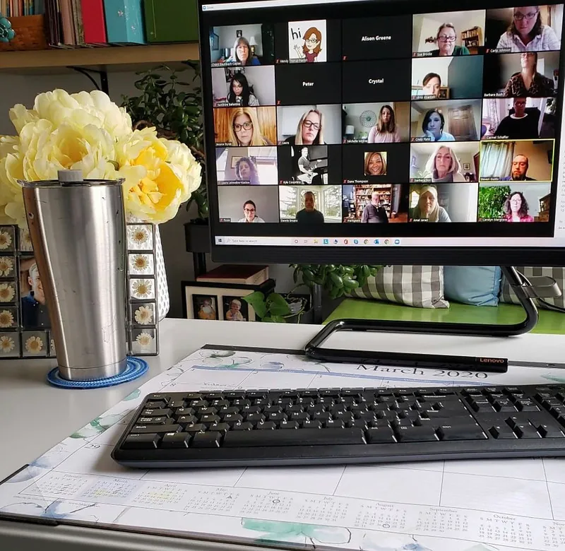 A computer monitor shows students in a virtual classroom during the 2020 pandemic. An insulated cup, flowers, and a keyboard appear near the monitor.