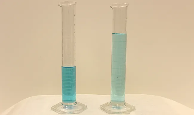Esta figura muestra dos cilindros graduados uno al lado del otro. El primero tiene aproximadamente la mitad de líquido azul que el segundo. El líquido azul es más oscuro en el primer cilindro que en el segundo.