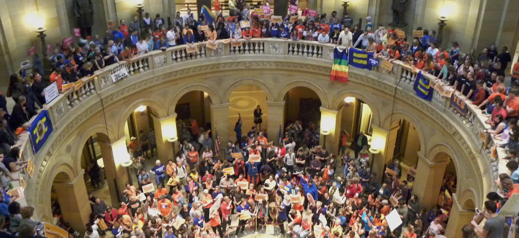 A large crowd of people pack two floors of a rotunda. Some hold rainbow flags or flags bearing an equals sign.