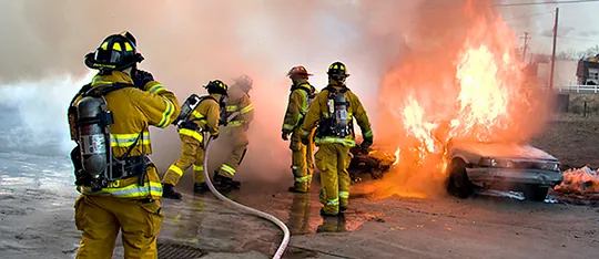 Photograph shows a group of firefighters in uniform using a hose to put out a fire that is consuming two cars.