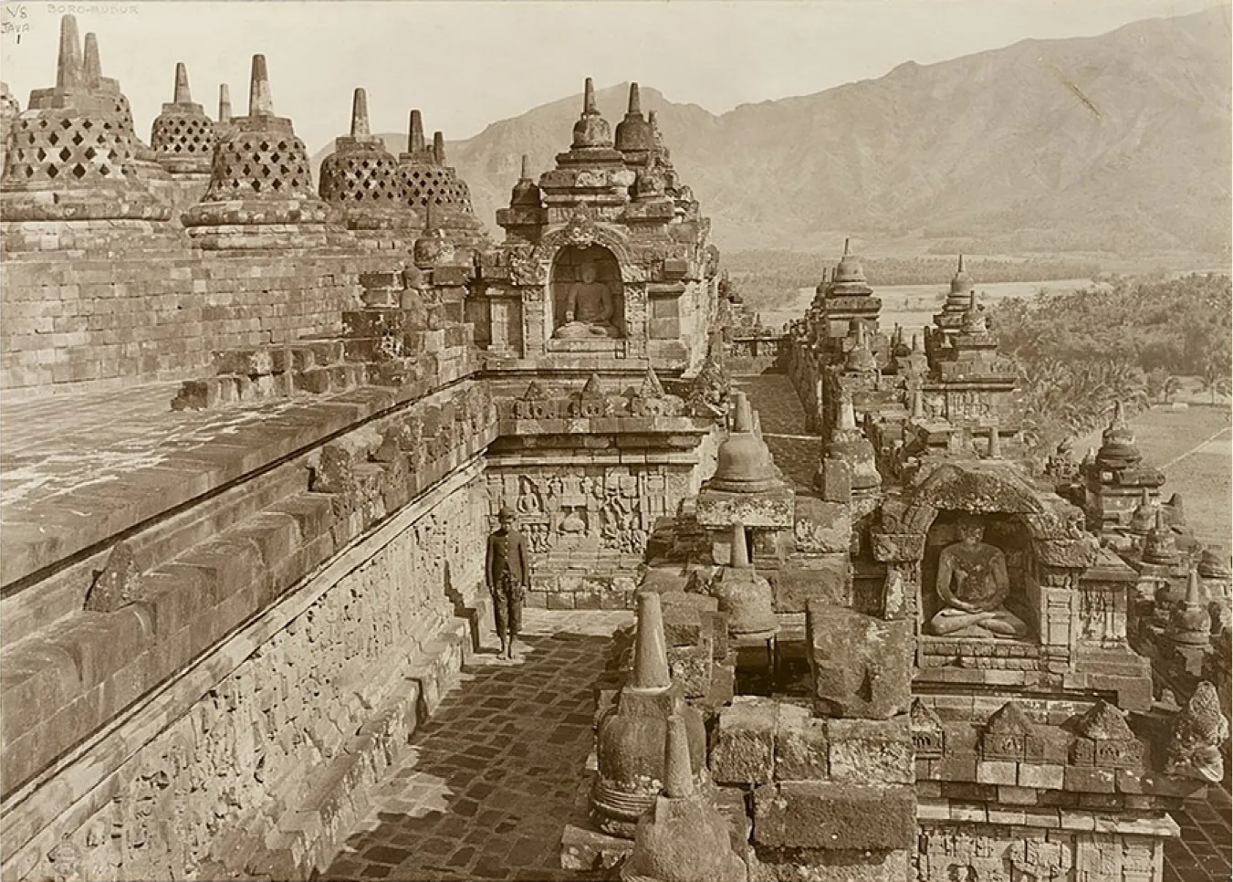An image of a sepia colored photograph is shown. The far right background shows tall mountains and areas of trees, water, and flat grassy areas show in front of the mountains on the right of the image. At the top left, eight mound-like structures (stupas) are shown sitting atop square bricked pedestals at varying levels. A thin vertical spire shows at the top and four rows of diamond shaped perforated openings line the mound. To the right, more solid stupas line the top of a structure with an arc shaped opening with a sitting statue inside. The archway is decorated with carvings. To the left of the structure on the same level as the stupas, a broken wall of bricks shows with the head of a statue showing. The next level down shows an uneven bricked walkway that extends out to the back of the massive structure. Lighter colored walls run along the walkway made of bricks and carvings show within those bricks. A figure stands along this wall under the sitting statue above in a dark buttoned jacket and pants that stop below the knee. He wears a dark cap and looks down. To the right of the walkway in the forefront, another sitting statue in an arched opening sits with legs crossed and hands resting in his lap. The statue is very worn and the stones above the arch are broken and missing pieces. Another walkway sits to the lower right of this structure with walls running along with carvings as described earlier. Along the right side, walls run with solid stupas lining the tops of varying levels.