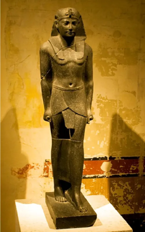 A stone statue is shown on a small pedestal along a peeling yellow and orange wall. The statue shows a man standing with his arms at his sides, hands made into fists, and his left foot in front of the right foot. He wears a long head covering that sits on his back and shoulders with a decorative object at his forehead. He is bare chested and wears a cloth around his waist. The statue is cracked in some places and forms shadows on the wall on both sides.