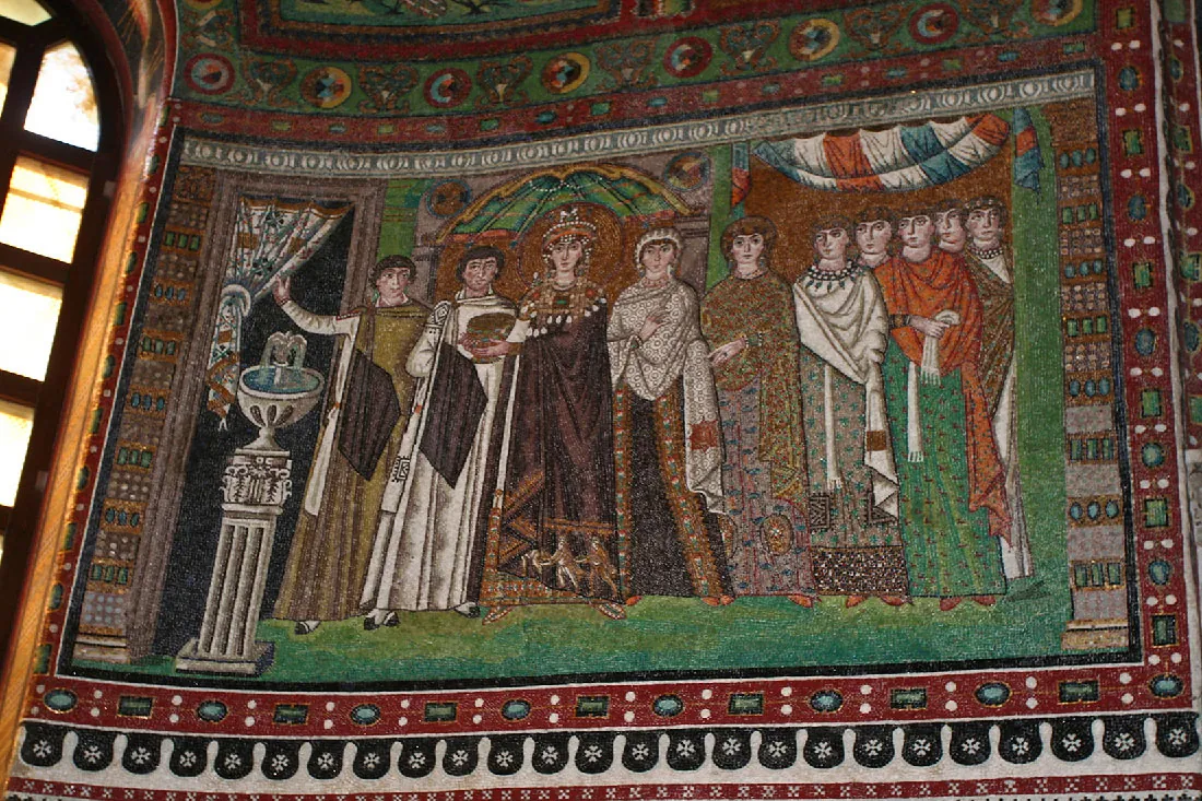A picture of a rectangle wall mosaic is shown. Ten people are shown standing in a line. They all wear some type of long, draped clothing. A white water fountain on a carved pedestal stands on the left side of the mosaic. The person farthest left is holding a red and white decorated drape open with his right hand and has short brown hair. He wears a green and brown long covering over a knee length, long sleeved, white shirt and has black tipped white shoes. The person next to him has a long white robe on with a brown square, short brown hair, black tipped white shoes, and accepts a dark, decorated chalice from the woman to his left. The woman wears a long, brown robe with jewels and tassels at the top and scenes stitched along the bottom. Her head is adorned with a jeweled crown and jeweled tassels hanging down on both sides. She wears dark shoes with circles on them. To her left is another woman in a dark, detailed robe and decorated, white shawl with a simple white headdress and red shoes. A woman is to her left in a dark, detailed long dress and large hairdo and earrings. Her left hand is shown wearing a large ring. Five men stand to her left in long detailed robes, red shoes, and short hair. Drapes are shown above their heads. The edges of the mosaic are laid with various shapes and decorations. A rounded window is seen at the left of the picture.