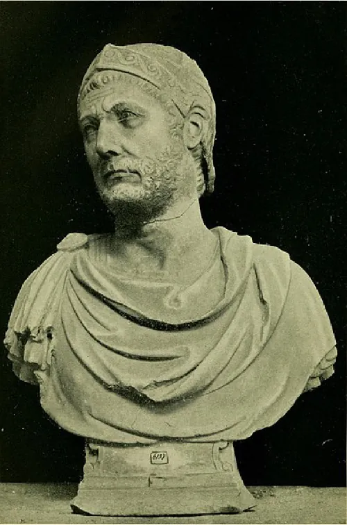 A picture of a gray stone bust is shown on a stone floor on a solid black background. The bust is of a man looking to his right with a sad expression. He has a beard, short curly hair, and a decorated helmet. There is a crack in the statue along the neck. He wears robes with a circular pin on his right shoulder.