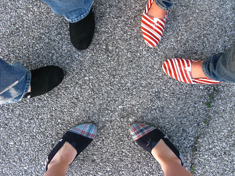 A photo of three pairs of feet wearing TOM’s shoes. The feet are positioned so they form a circle. One person is wearing TOMs with a striped pattern; another person is wearing plaid patterned TOMs, and the last person is wearing solid black TOMs.