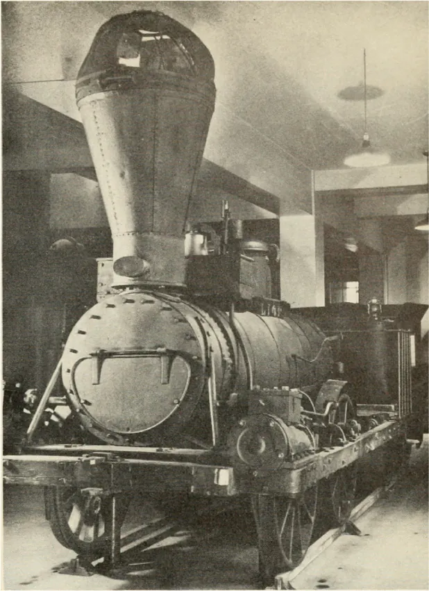 A photograph of a steam locomotive in a museum. The locomotive body is a large cylinder and it has a tall chimney stack emerging from the front of the body.