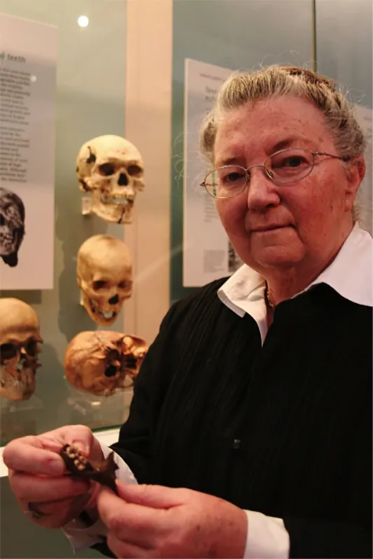 Woman with white hair and wire rim glasses standing in front of a display case filled with skulls.