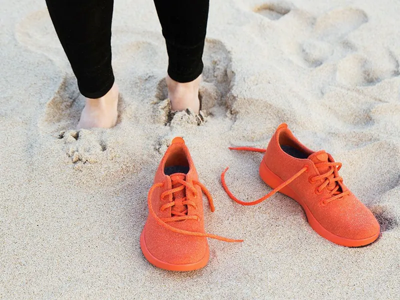 A pair of untied shoes are on top of the sand. Behind the shoes, a person stands with their feet buried in the sand.