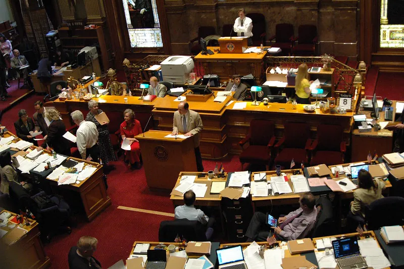 In a busy state senate chamber, professionally dressed legislators, some working at laptops, sit behind desks covered in papers or stand conferring with their colleagues while a person stands at a podium facing them.