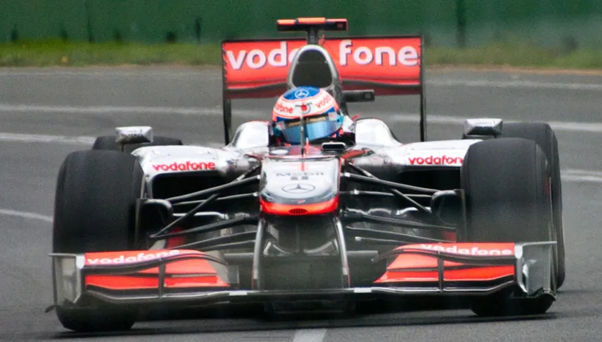 The figure shows, from front, a red and silver coloured Formula One car turning through a curve in a race on the Melbourne Grand Prix track, with the driver in seat.