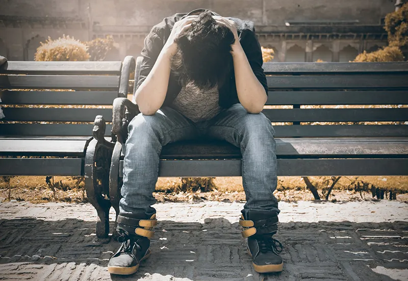 An outdoor photo shows a depressed young man sitting on a bench in closed position with his head down.
