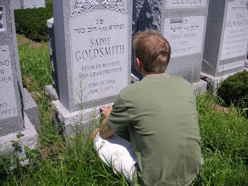 A young man in a green T-shirt and white shorts is shown sitting in the grass in front of a gravestone.