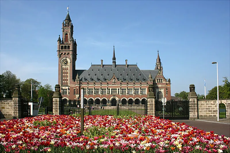 A large brick building is surrounded by brick walls and iron gates. A tall clock tower rises on the left side of the building. A field of colorful flowers blooms in front of the building and gates.