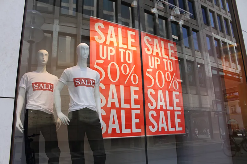 A retail window shows two mannequins wearing shirts that say sale. There are two signs in the window that say sale up to 50% sale sale.