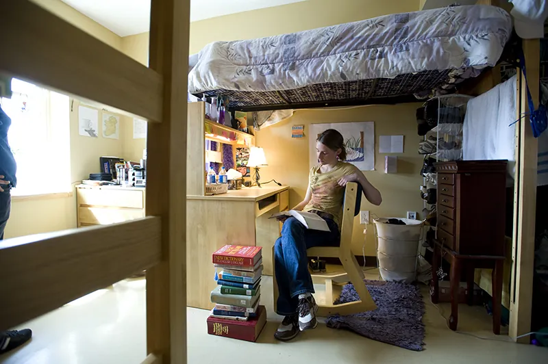 A female student wearing jeans and a t-shirt reading at a desk and study area underneath an elevated bed, with more books stacked on the floor.