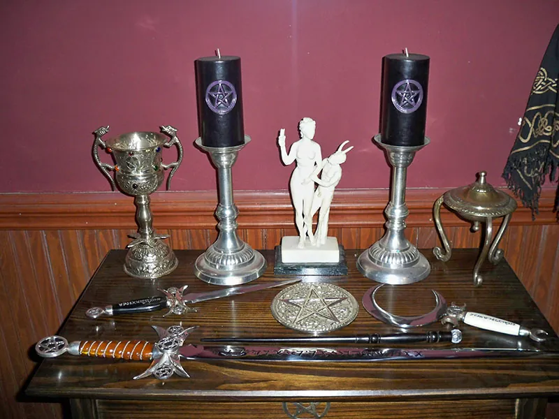 A small wooden table with various objects arranged on top of it. Among the objects are a statue of two human figures, two tall candle holders with silver bases, a knife and a sword with elaborate hilts, and a silver chalice.