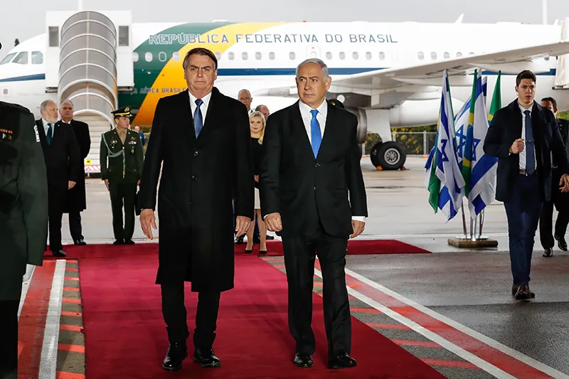 Brazilian President Jair Bolsonaro and Israeli Prime Minister Benjamin Netanyahu walk down a red carpet at an airport. A Brazillian plane is parked directly behind them. People in the background wear suits and military uniforms. Five or six Israeli and Brazilian flags are present to the right of the red carpet.