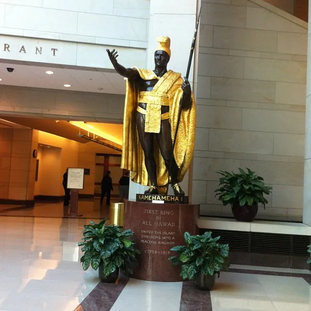A statue of a man standing upright with his hand extended in an oratory gesture. He holds a staff in the other hand. The statue is wearing a gold cloak and hat and stands in a contemporary-looking indoor space.