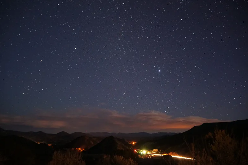 Evening sky, with stars dominating the top portion of the image, low clouds on the horizon, and mountainous terrain at the bottom. A spot of electric lights creates a bright glow.
