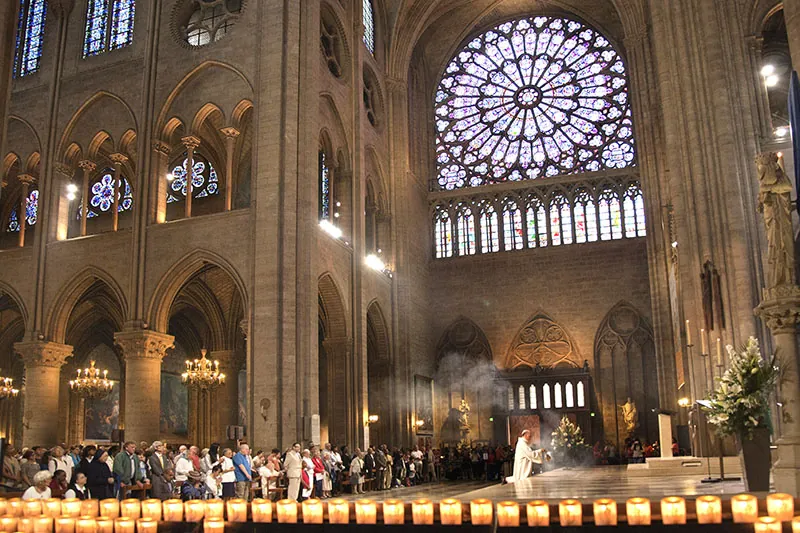 Interior image of the Notre Dame cathedral during a church service. Two tiers of gothic arches are visible and above these are stained glass windows in the shape of medallions. The human figures in the image are very small, emphasizing the size of the cathedral.