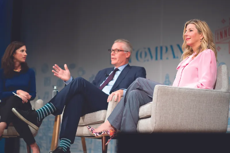 Photo of three people sitting on a stage, talking, with Sara Blakely on the right.