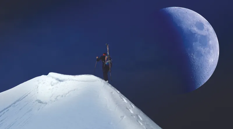This image shows a person hiking up a snow-covered mountain. The moon is half-full and off to the right of the mountain and the hiker. It appears very large in the sky and very close to the hiker.