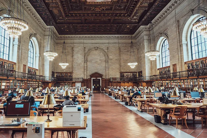 Large, spacious room with very high ceilings. Books shelves line the walls. Rows of tables, dotted with small reading lamps, stretch along the sides, with a wide aisle between them.