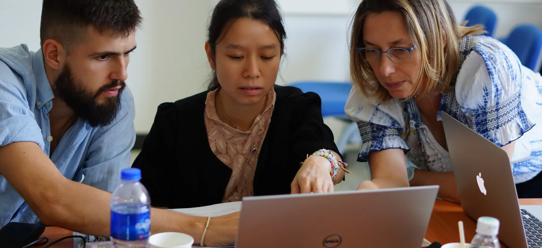 Three people are collaborating around two laptop computers. Water bottles and cups are also visible on the table.