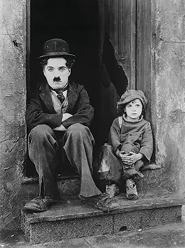 Charlie Chaplin is shown sitting in a doorway with his arms folded, accompanied by a small, shabbily dressed child.