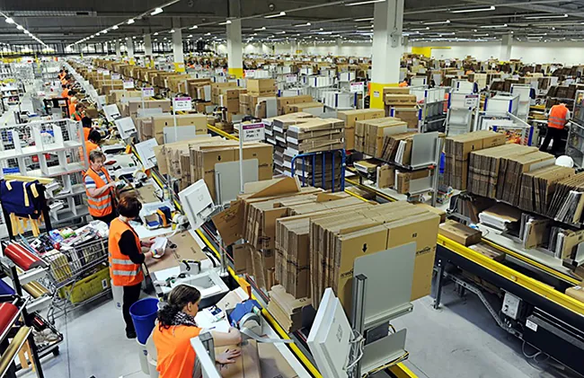 A photo shows a view of an Amazon logistics center with employees packing parcels to be shipped.