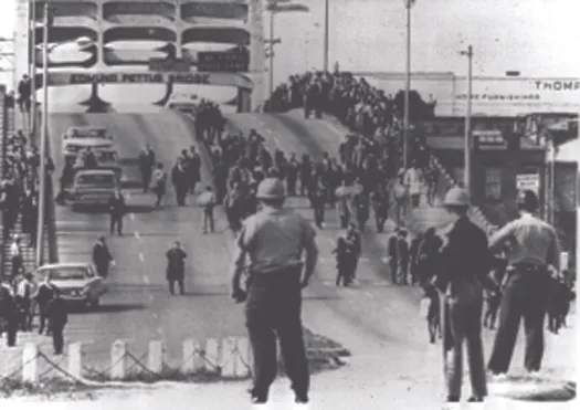 An image of a bridge. On the right of the overpass are several people marching in a large crowd. In the foreground are uniformed people watching the marchers.