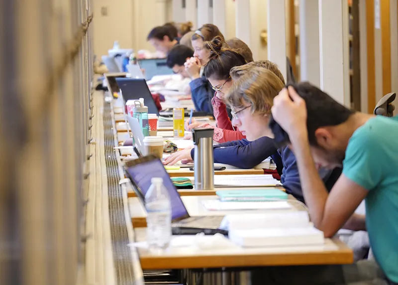 A group of students sit at a long table reading and working at computers. They have various books, papers, and notebooks.