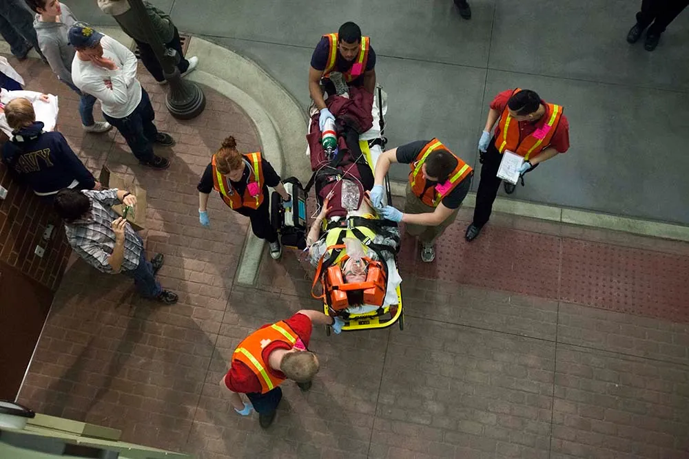 An overhead view of a paramedics team of five moving a patient secured on a stretcher through a public place. People stand beside watching them walk away.