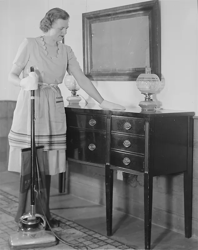 An American woman is dusting a sideboard. She is holding an electric vacuum cleaner in the other hand.