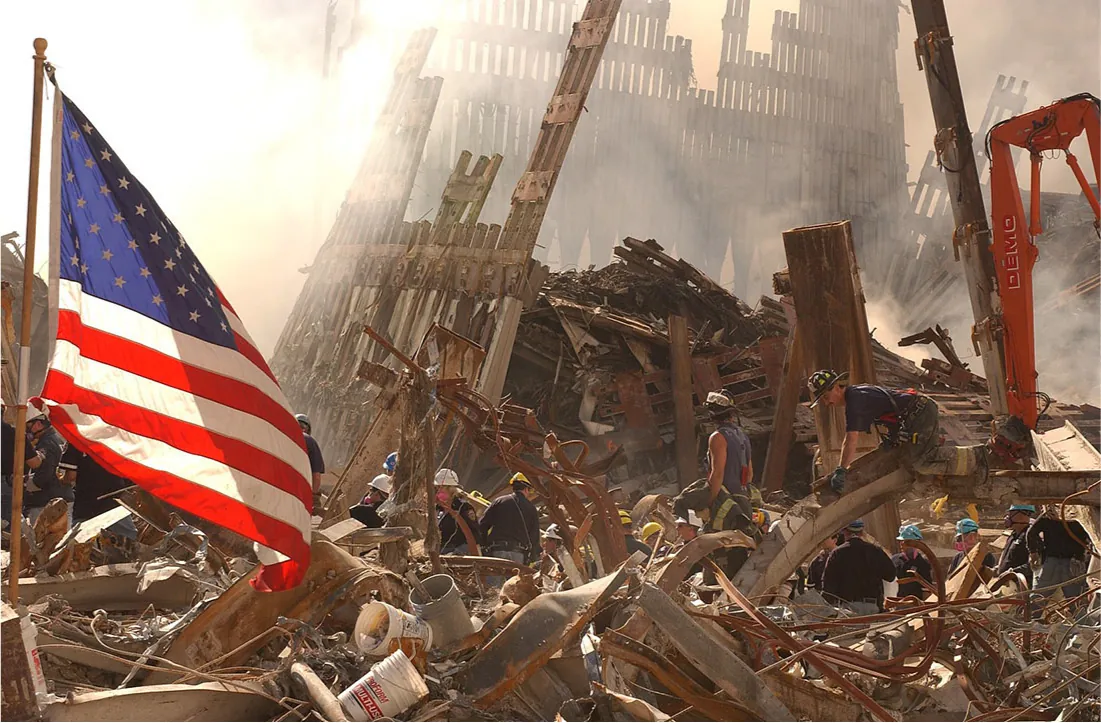 A picture of a crumbled building is shown. Firemen and other people in hard hats are walking amid the twisted and burnt rubble, moving objects and picking things up. Two tall gridded walls are seen on the left side and in the back bent against the rubble on the ground. A large red crane is shown at the right and the American flag with red and white stripes and white stars on a blue background in the left corner is shown flying in the left side of the picture next to buckets strewn about.