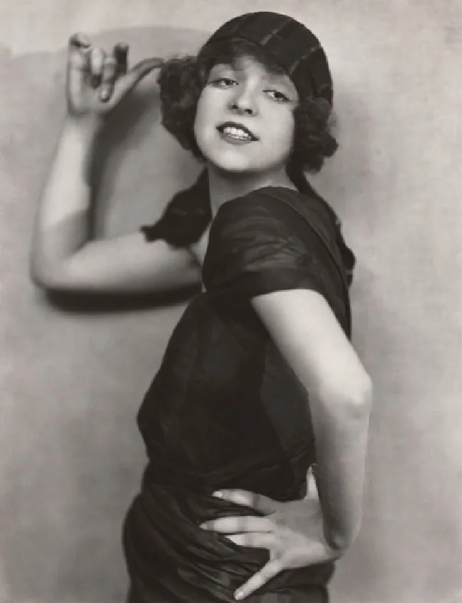 A woman wears a dark dress and striped hat. She smiles and stairs into the distance. Her left hand is on her hip and her right hand is beside her head. She appears to be intentionally posing for the camera.