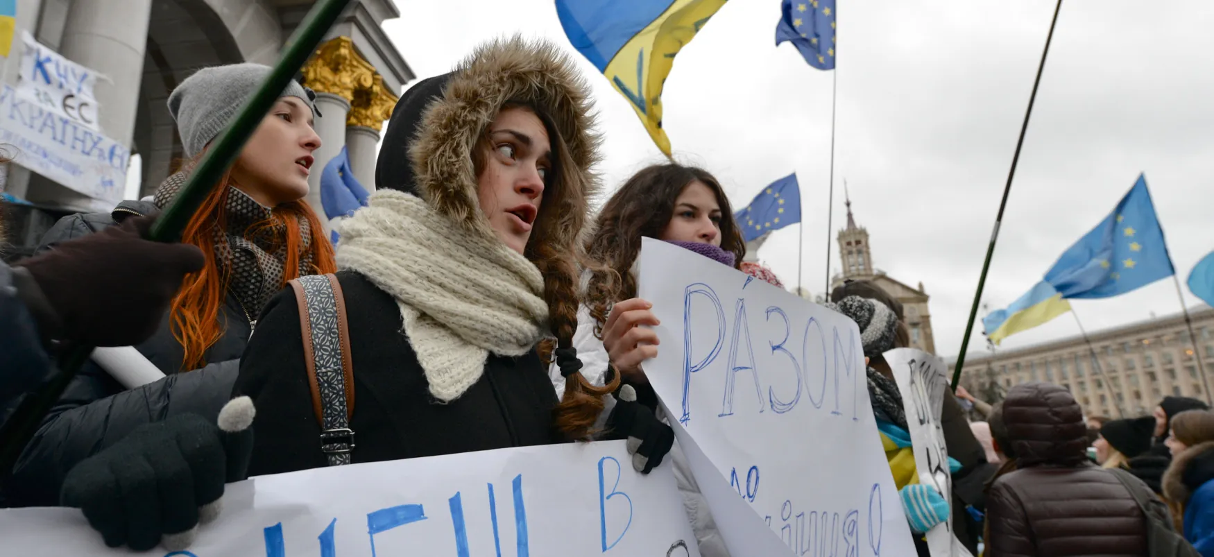 Three young people hold signs at a protest. Other protesters,  some carrying EU flags, stand in the background.