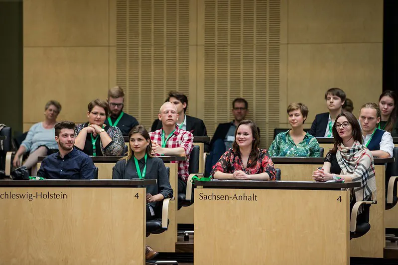 Young adult student journalists wearing lanyards sit facing forward in tiered rows of two-person desks.