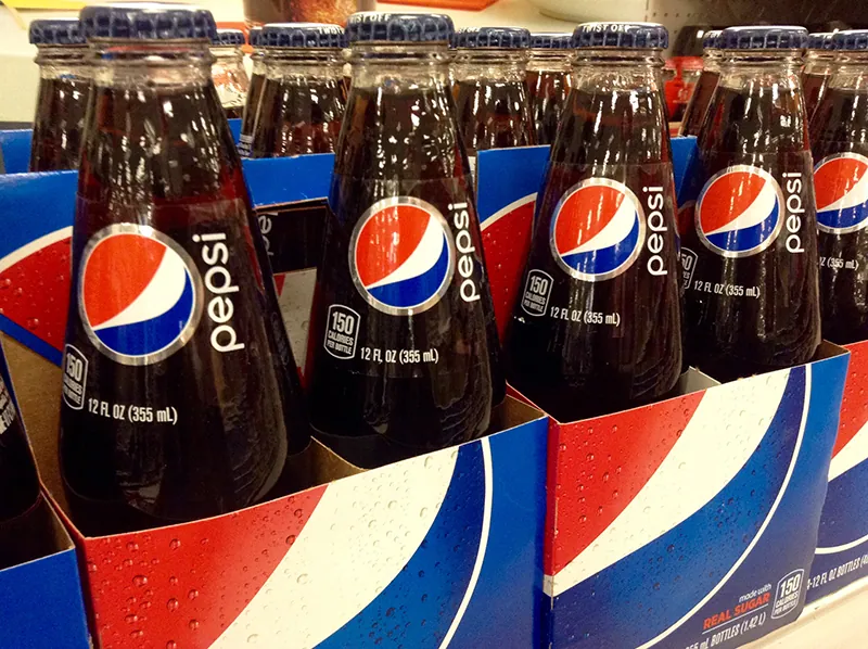 Four packs of Pepsi in glass bottles are lined up in a row. The Pepsi logo is on both the bottle and the cardboard packaging.