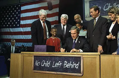 A photograph shows President Bush signing the No Child Left Behind Act at a large desk, surrounded by U.S. officials and several children. On the desk hangs a chalkboard that reads “No Child Left Behind.”