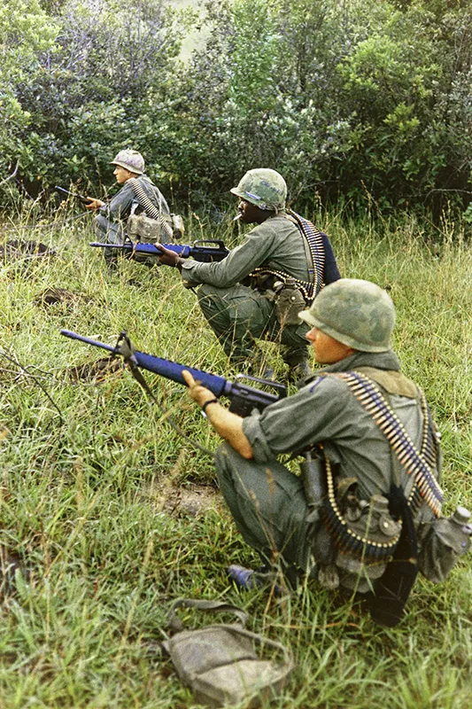 Three soldiers in uniform and combat helmets kneel in a field holding rifles.